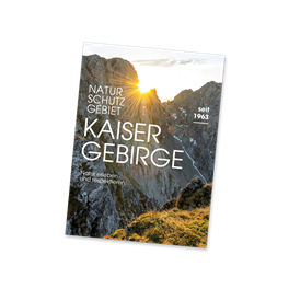 Ein Buch über das Kaisergebirge mit majestätischen Bergen im Hintergrund. Der Sonnenuntergang verleiht der Szene eine warme Atmosphäre.