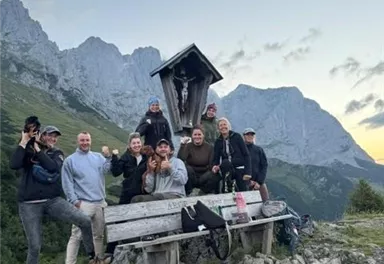 Eine Gruppe von zehn Personen steht fröhlich auf einem Berg. Im Hintergrund sind beeindruckende Felsen und ein klarer Himmel zu sehen.