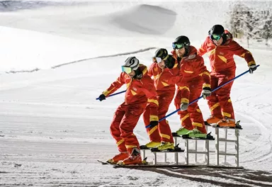 A group of skiers in orange clothing are skiing on a slope. They are standing on a construction made of ladders and showcasing a spectacular ski action.