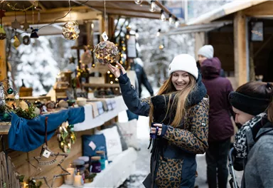 Eine fröhliche junge Frau auf einem Weihnachtsmarkt hält eine dekorative Glaslaterne. Im Hintergrund sind Stände mit festlicher Beleuchtung und anderen Besuchern zu sehen.