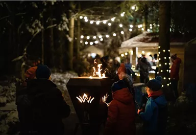 Eine winterliche Szene mit einer Feuerstelle, umgeben von Menschen. Im Hintergrund sind Lichterketten, die eine gemütliche Atmosphäre schaffen.