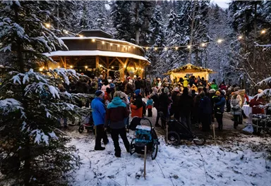 Eine festliche Zusammenkunft im Schnee mit vielen Menschen, die sich um festlich beleuchtete Holzhütten versammeln. Die winterliche Landschaft ist von Tannenbäumen umgeben.