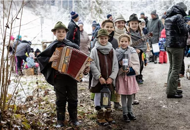 Eine Gruppe von Kindern in traditioneller Kleidung steht auf einem Weg im Schnee. Einige von ihnen halten Musikinstrumente und lächeln in die Kamera.