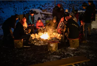 Eine Gruppe von Menschen sitzt um ein Lagerfeuer in einer winterlichen Umgebung. Die Atmosphäre ist gemütlich und einladend.