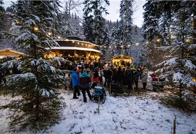 Eine winterliche Festveranstaltung im Wald mit vielen Menschen. Die Umgebung ist mit Schnee bedeckt und das Licht der Veranstaltung schafft eine gemütliche Atmosphäre.