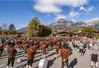 Eine Gruppe von Menschen in traditioneller Tracht versammelt sich auf einem Platz. Im Vordergrund geht ein Mann mit einer Schärpe über eine Holzlatte.