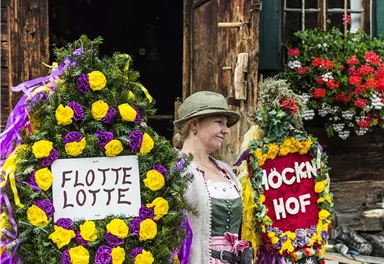 Eine Frau in traditioneller Tracht steht vor einem Holzhaus und trägt zwei festlich geschmückte Kränze. Die Kränze sind mit bunten Blumen dekoriert und tragen die Schriftzüge "FLOTTEN LOTTE" und "HÖCKEN HOF".