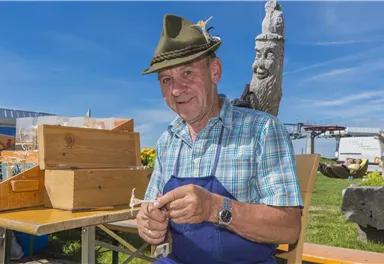 Ein lächelnder Mann trägt einen traditionellen Hut und arbeitet an einem Tisch im Freien. Hinter ihm sind Holzkisten und eine grüne Wiese zu sehen.