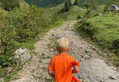 Ein Junge in einem orangefarbenen Shirt geht einen schmalen Pfad in einer bergigen Landschaft. Im Hintergrund sind grüne Hügel und teilweise bewölkter Himmel zu sehen.