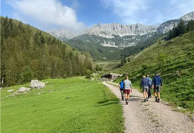 Eine Gruppe von Menschen wandert auf einem Pfad durch eine grüne Wiese in den Bergen. Im Hintergrund sind hohe, bewaldete Hügel und ein klarer Himmel zu sehen.
