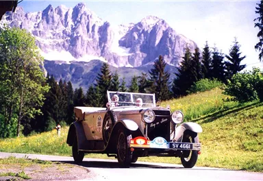 A classic car drives along a picturesque road in the mountains. The surroundings are green with tall trees and impressive mountain peaks in the background.
