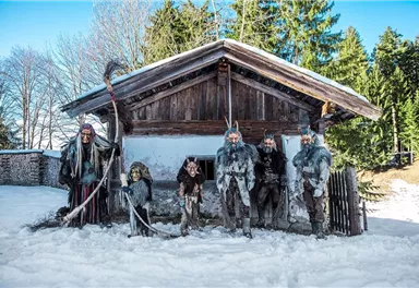Eine Gruppe von Personen in traditionellen Kostümen steht vor einem alten Holzhaus im Schnee. Im Hintergrund sind Bäume und blauer Himmel zu sehen.