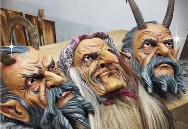 Drei gruselige Masken mit Hörnern und langen Bärten liegen auf einem Tisch. Die Masken zeigen furchterregende Gesichter mit unterschiedlichen Ausdrücken.