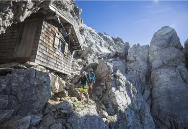 Eine Berghütte steht zwischen felsigen Klippen. Zwei Wanderer erkunden die Umgebung unter einem klaren blauen Himmel.