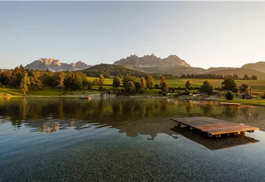 A calm lake with a wooden jetty and mountains in the background. The landscape is green and the atmosphere is peaceful.