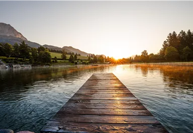 A calm lake with a wooden plank leading into the water. In the background, the mountains are visible and the sun rises in the morning air.