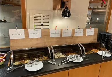 Ein Buffet mit verschiedenen Speisen in warmen Behältern. Darüber hängen Beschilderungen mit den Namen der Gerichte.