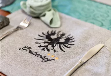 Ein eleganter Tischdecken mit dem Wappen und dem Namen "Grudenhof". Das Design ist schlicht und einladend, mit Besteck ordentlich angeordnet.