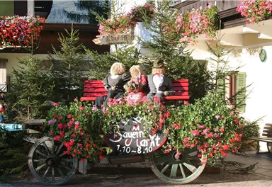 A decorated wooden cart with colorful flowers and three dolls sits on a bench. In the background, green fir trees and a traditional building can be seen.