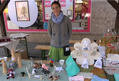 A woman stands at a market stall with various handmade items. On the table, toys, bottles, and other creative products are displayed.