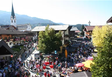 A bustling marketplace with many people and colorful stalls. In the background, traditional buildings and a church tower can be seen.