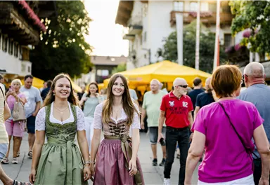 Two women in traditional dirndl dresses are happily walking through a busy street. Stalls and many people can be seen in the background.