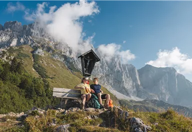 Zwei Personen sitzen auf einer Bank in der Berglandschaft. Im Hintergrund sind hohe Berge und ein klarer Himmel sichtbar.