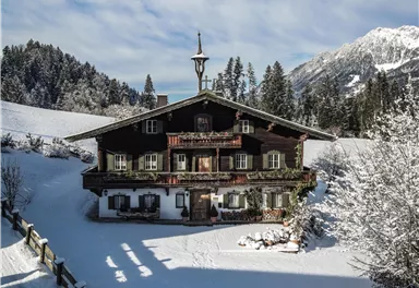 Ein traditionelles Chalet im Schnee, umgeben von Tannenbäumen. Der Himmel ist blau und wolkig, was eine idyllische Winterlandschaft schafft.