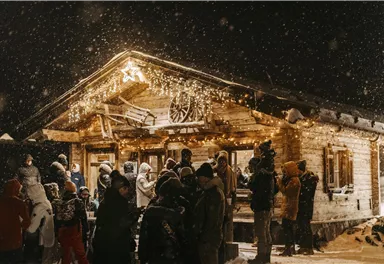 Eine gemütliche Hütte in der Nacht, umgeben von starkem Schneefall. Viele Menschen stehen draußen und genießen die festliche Beleuchtung.