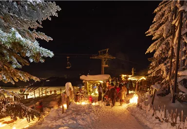 Eine verschneite Straße bei Nacht, beleuchtet von Fackeln und umgeben von schneebedeckten Bäumen. Im Hintergrund sind Menschen und eine Gondelbahn zu sehen.
