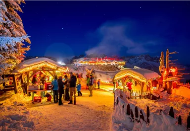 Ein festlicher Weihnachtsmarkt in einer verschneiten Landschaft. Die Stände sind mit Lichtern geschmückt und Menschen genießen die Atmosphäre bei Nacht.