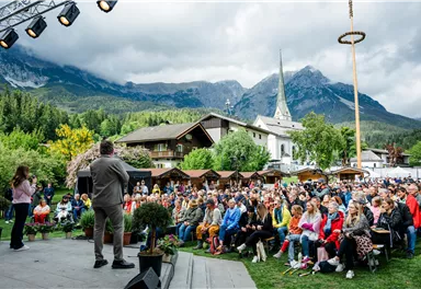 A large audience is sitting outdoors in front of a stage while a speaker is speaking. In the background, mountains and a church can be seen, creating a picturesque setting.