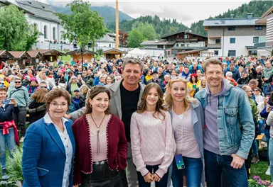 A group of five people is smiling in front of a large crowd. In the background, mountains and buildings can be seen.