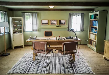 Ein gemütliches Arbeitszimmer mit einem großen Holzschreibtisch und zwei Stühlen. Die Wände sind in einem sanften Grün gestrichen, und es gibt Bücherregale sowie Fenster mit Vorhängen.