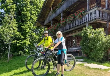 Zwei Radfahrer stehen vor einem traditionellen Holzhaus in der Natur. Im Hintergrund sind grüne Bäume und eine schöne Aussicht zu sehen.