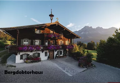 A traditional wood-clad house with colorful flower boxes. In the background, majestic mountains and a green landscape can be seen.