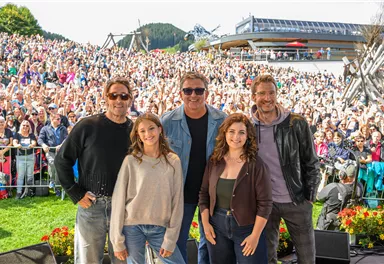 Five people are standing smiling in front of a large crowd. In the background, mountains and a building can be seen.