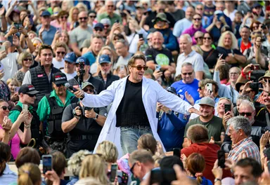 A man in a white lab coat stands at the center of a large crowd. The spectators are applauding and taking photos.