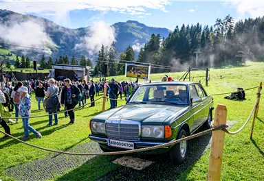 A classic car is standing on a green field. In the background, people and mountains can be seen, and the atmosphere feels festive.