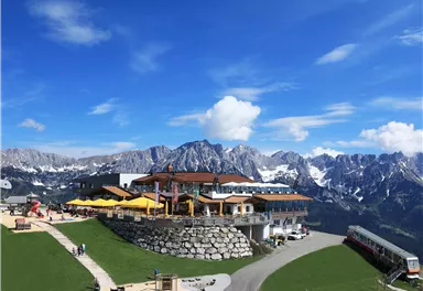 Eine malerische Berglandschaft mit einem Restaurant und Sonnenschirmen. Im Hintergrund sind beeindruckende Berge und ein blauer Himmel zu sehen.