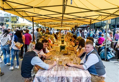 A lively street event with long tables under a yellow tent. People sit together, enjoying food and drinks, while a large crowd stands in the background.