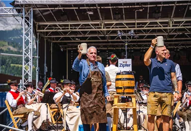 Zwei Männer heben Bierkrüge auf einer Bühne während einer Feier. Im Hintergrund sind Musiker in traditionellen Kostümen zu sehen.
