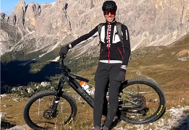 A mountain biker is standing with his bike in the Dolomites. In the background, high mountains and a clear blue sky are visible.