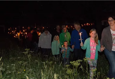 A group of people is walking at night, all with candles in hand. Trees and a meadow can be seen in the background.