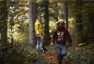 Ein Junge läuft fröhlich auf einem Waldweg, während eine Frau in einem gelben Mantel ihm folgt. Die Umgebung ist herbstlich mit buntem Laub und hohen Bäumen.