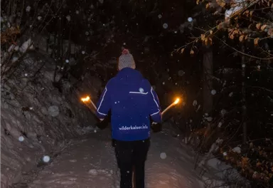 Eine Person läuft mit Fackeln auf einem verschneiten Weg in der Nacht. Es fällt Schnee und die Umgebung ist dunkel und ruhig.