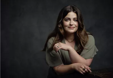 A smiling woman with long, brown hair is relaxing against an old chest. The background is dark and creates an elegant atmosphere.