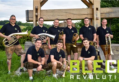 A group of musicians stands in front of a wooden structure holding various wind instruments. They are wearing black T-shirts and showing cheerful faces.