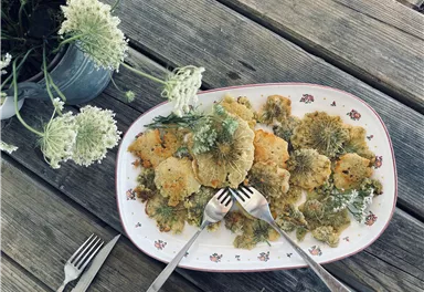 A plate of fried cauliflower, garnished with fresh herbs. In the background, flowers and cutlery can be seen on a wooden table.