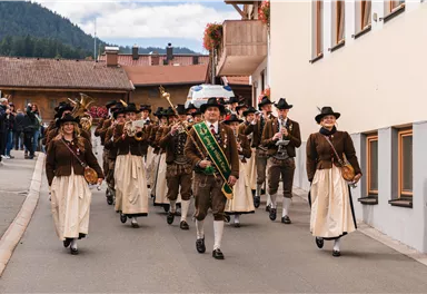 Eine traditionelle Trachtenparade mit Menschen in festlichen Kleidern läuft durch eine Straße. Im Vordergrund führt ein Mann mit einer Fahne die Gruppe an.